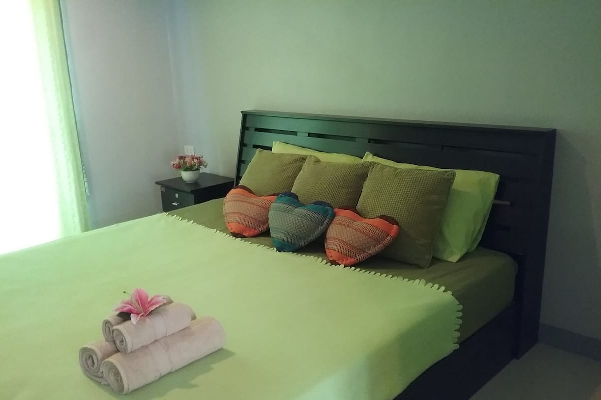 Una cama bien hecha con cubrecama verde, varios almohadones y toallas enrolladas adornadas con flores.