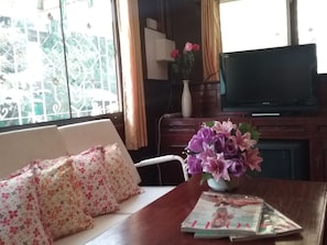 A living room with a sofa, a table with a vase of flowers, a flat-screen TV, and a window with a view of greenery.
