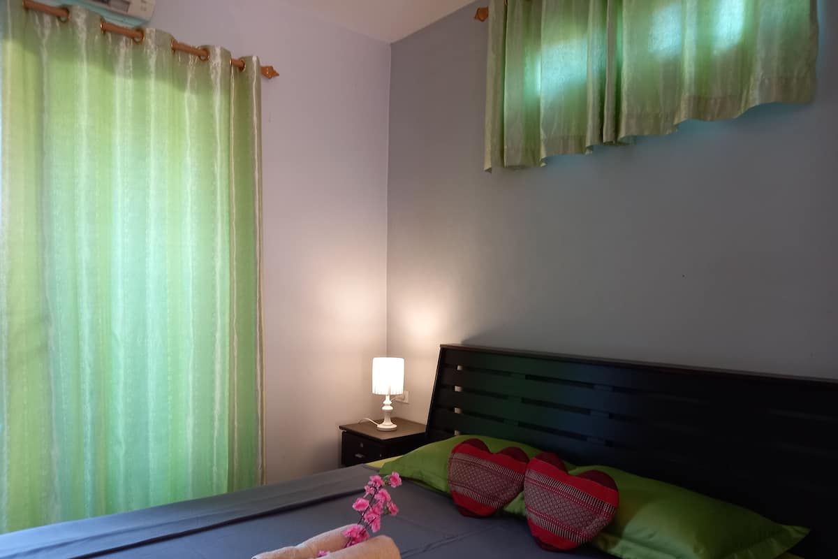 Una cama bien hecha con toallas enrolladas y almohadas con forma de corazón, una mesita de noche con lámpara y cortinas verdes.