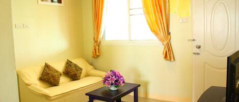 A room with a beige sofa, a small table with a vase of flowers, a window with curtains, and a door.