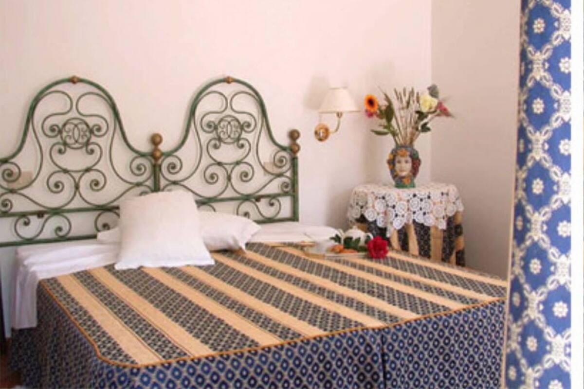A neatly made bed with a patterned bedspread, a decorative headboard, a small table with a vase of flowers, and a wall-mounted lamp.