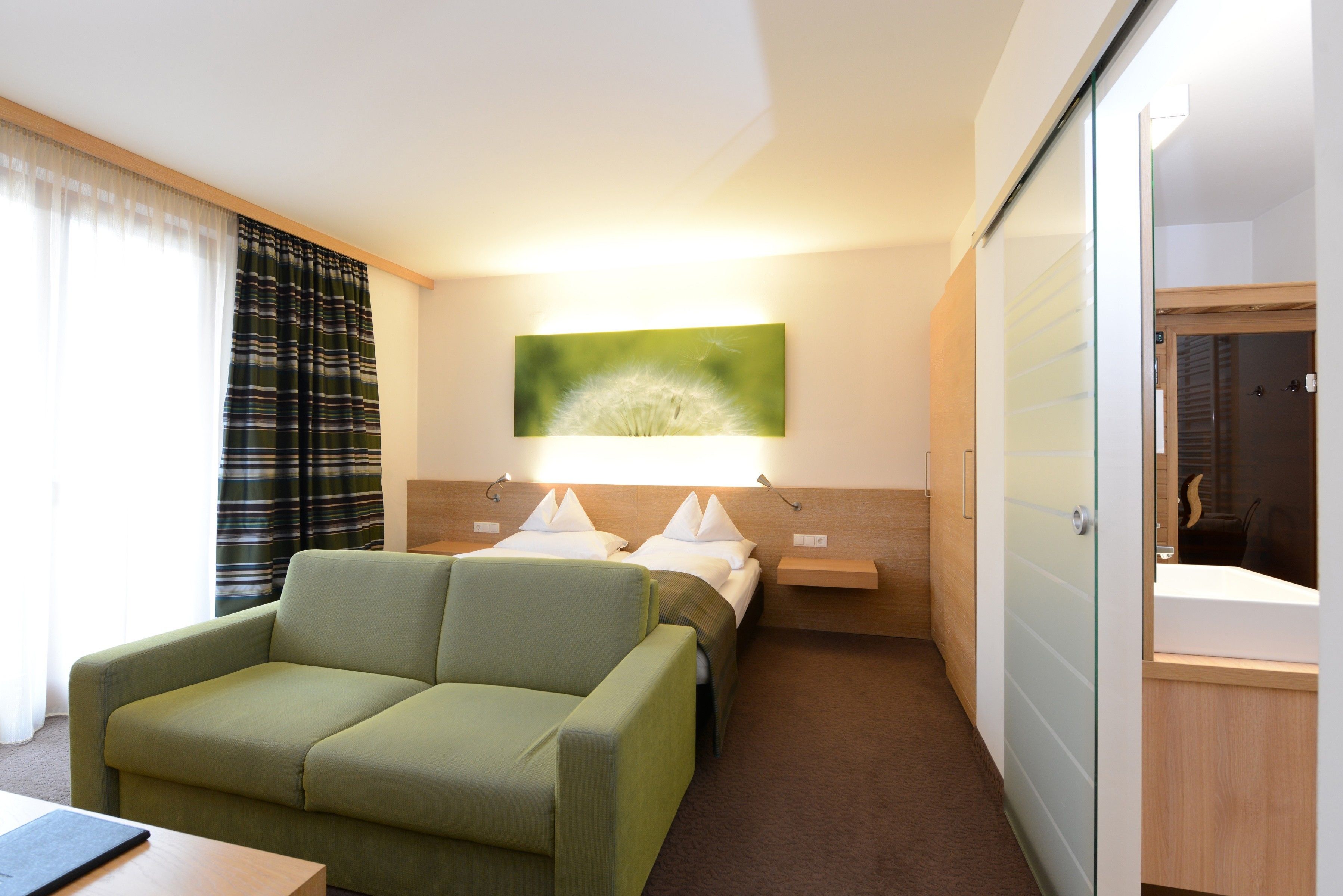 Habitación de hotel con un sofá verde, una cama con almohadas blancas, una lámpara de pared y un cuadro de un diente de león.