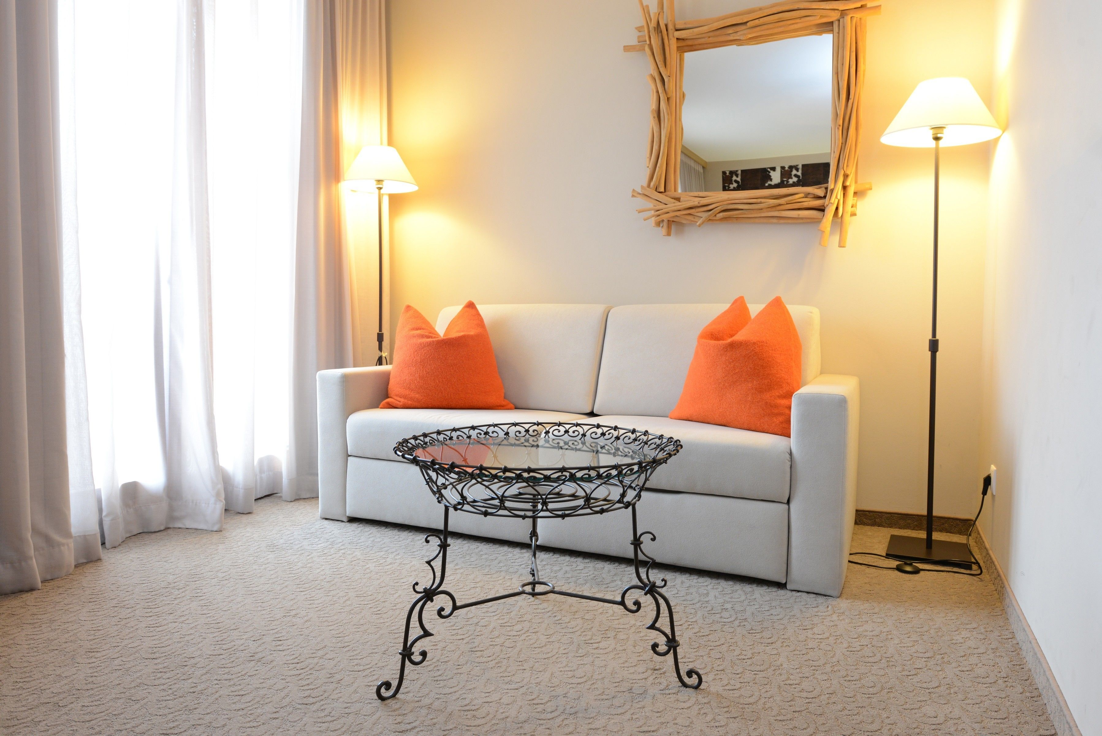 A living room with a white sofa, orange cushions, a glass coffee table with a metal base, and two floor lamps.