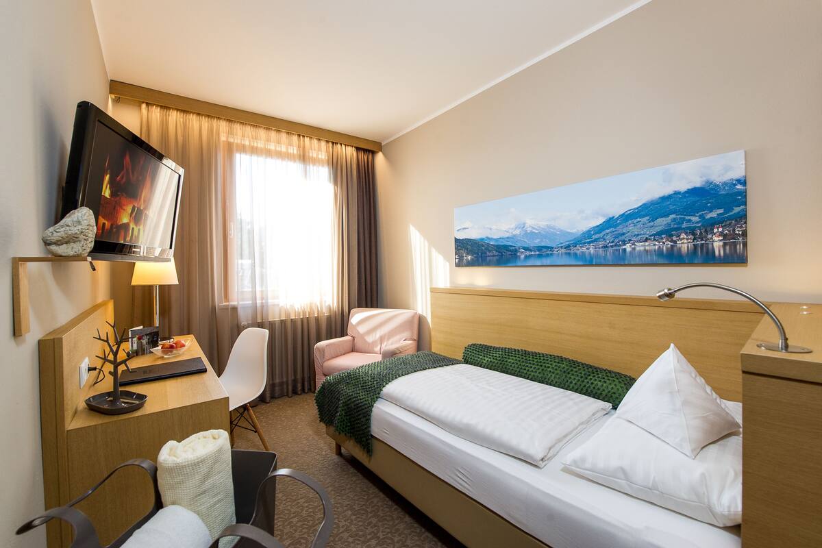 Habitación de hotel con cama, escritorio con ordenador, silla, televisor y cuadro de un paisaje montañoso.