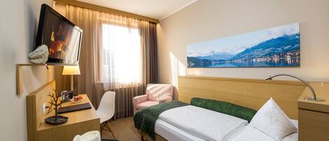 Habitación de hotel con cama, escritorio con ordenador, silla, televisor y cuadro de un paisaje montañoso.