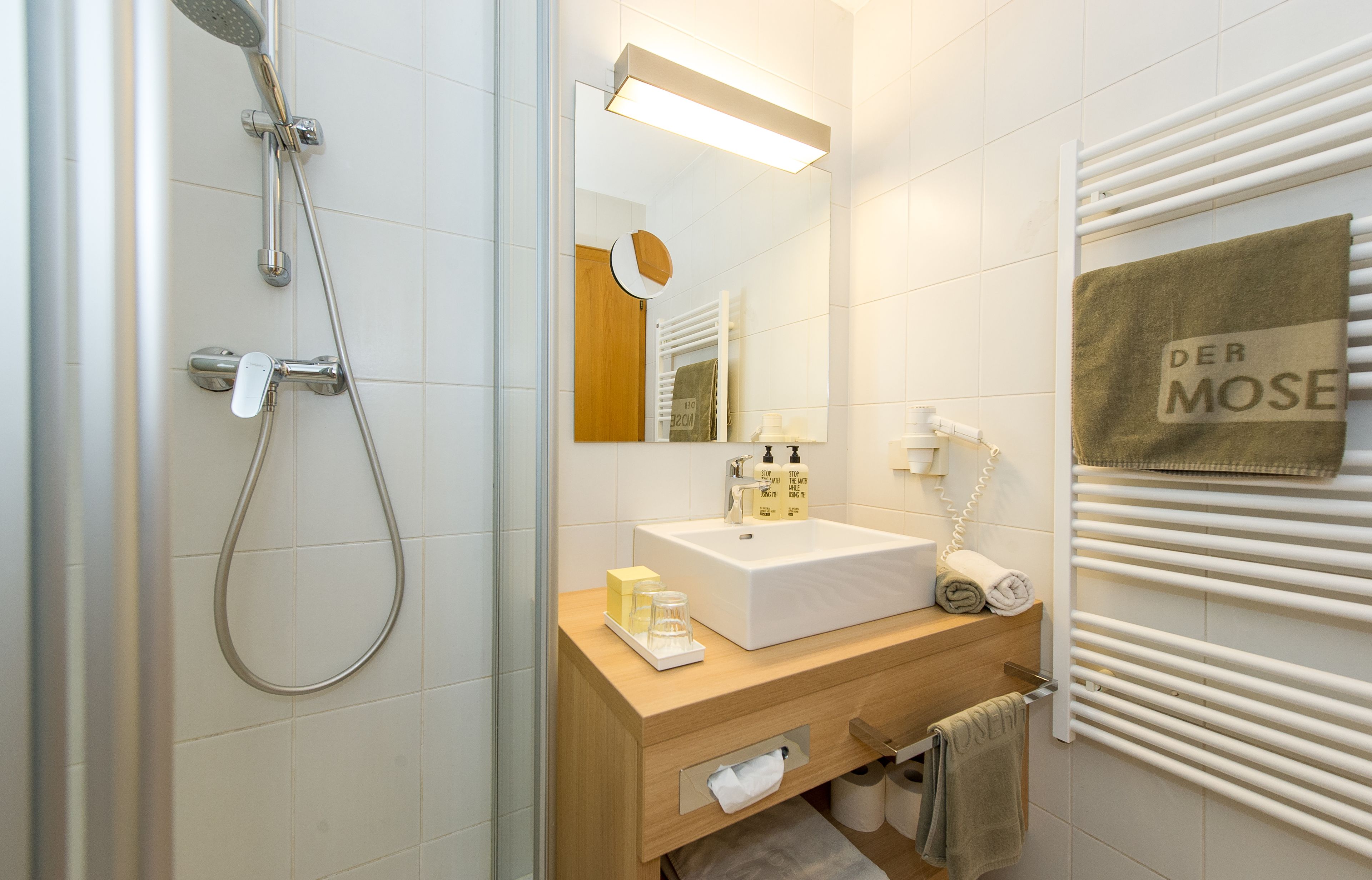 A modern bathroom with a shower, sink, mirror, towel rack, and a towel with "DER MOSE" on it.