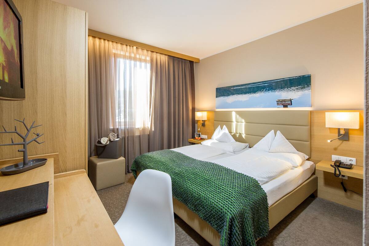 Habitación de hotel con cama, escritorio, silla, mesita de noche, lámpara y un cuadro en la pared.