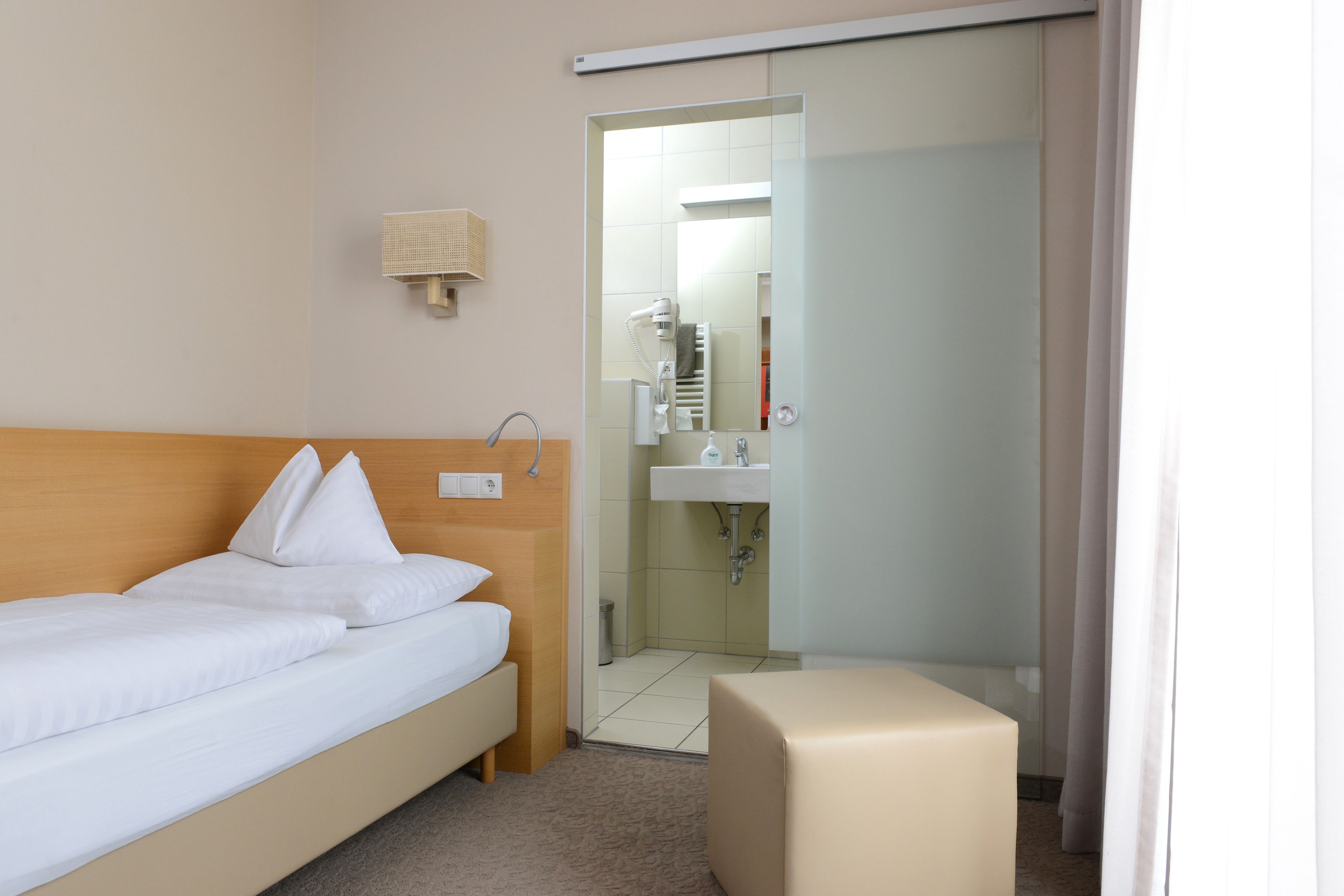 A hotel room with a bed, a bedside table, a small stool, a bathroom with a sink and mirror, and a shower.