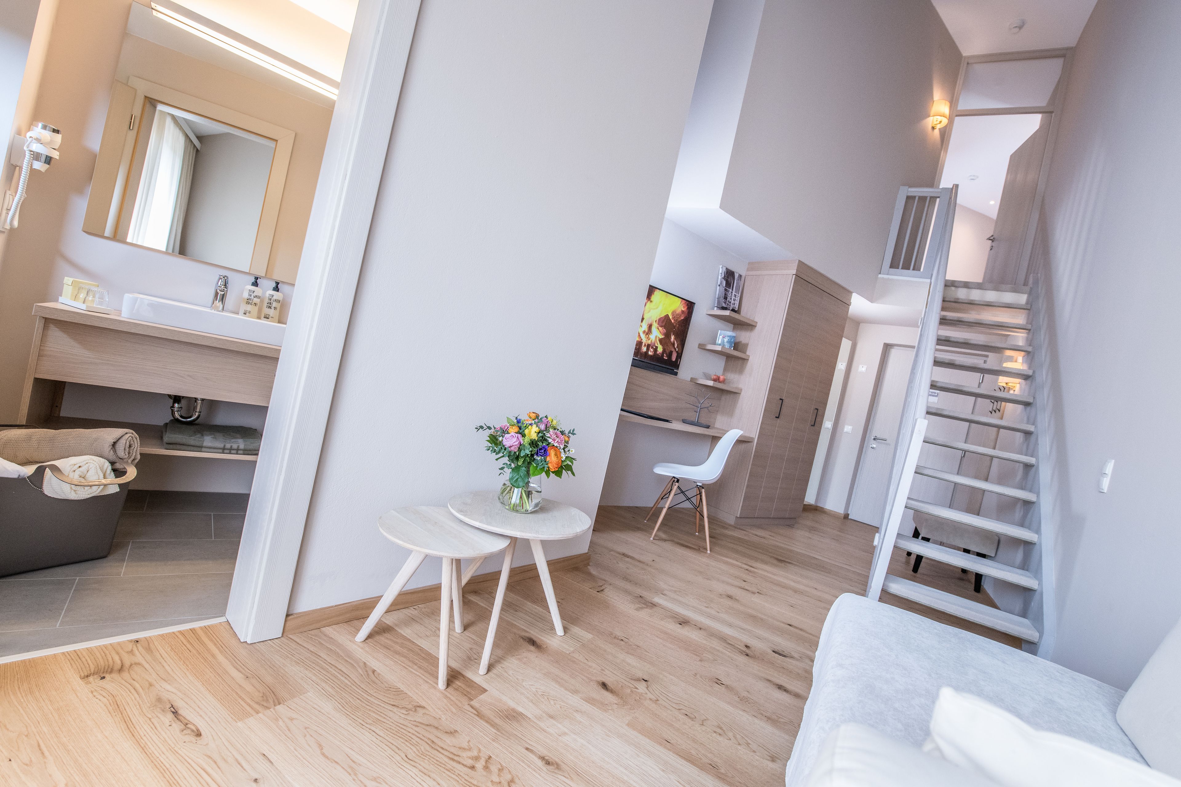 Un interior moderno con suelo de madera, un sofá blanco, una mesita redonda con un jarrón de flores, una silla blanca, una escalera y un baño con espejo y lavamanos.