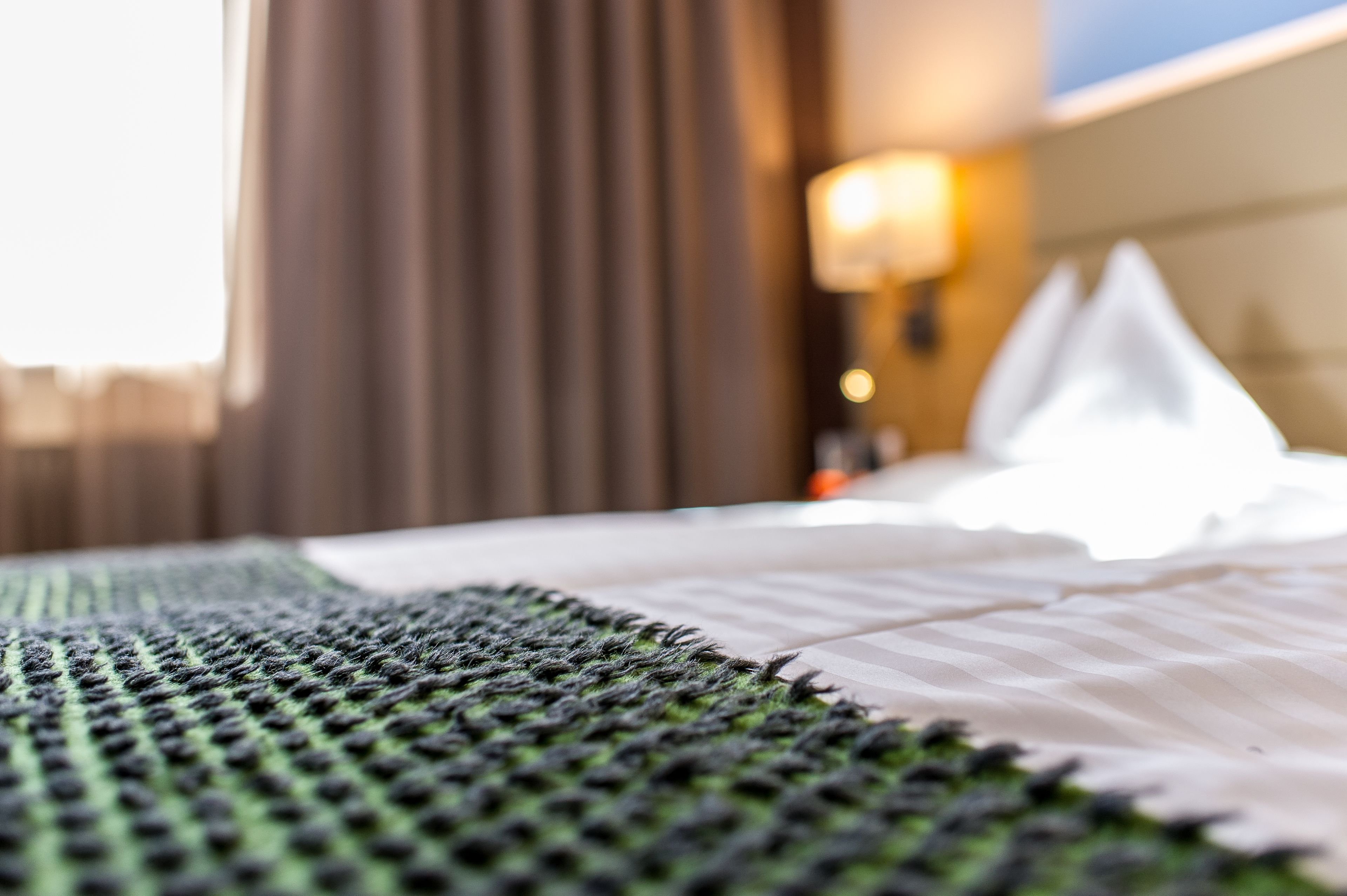 A close-up of a bed with a green and black textured blanket, a white pillow, and a blurred background with a lamp and curtains.