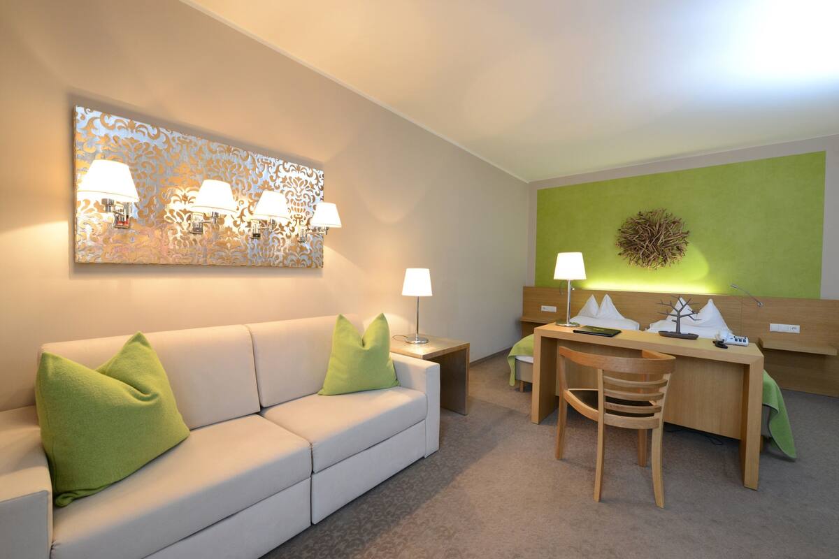 Habitación de hotel moderna con sofá blanco, almohadas verdes, escritorio con silla y un adorno de pared.