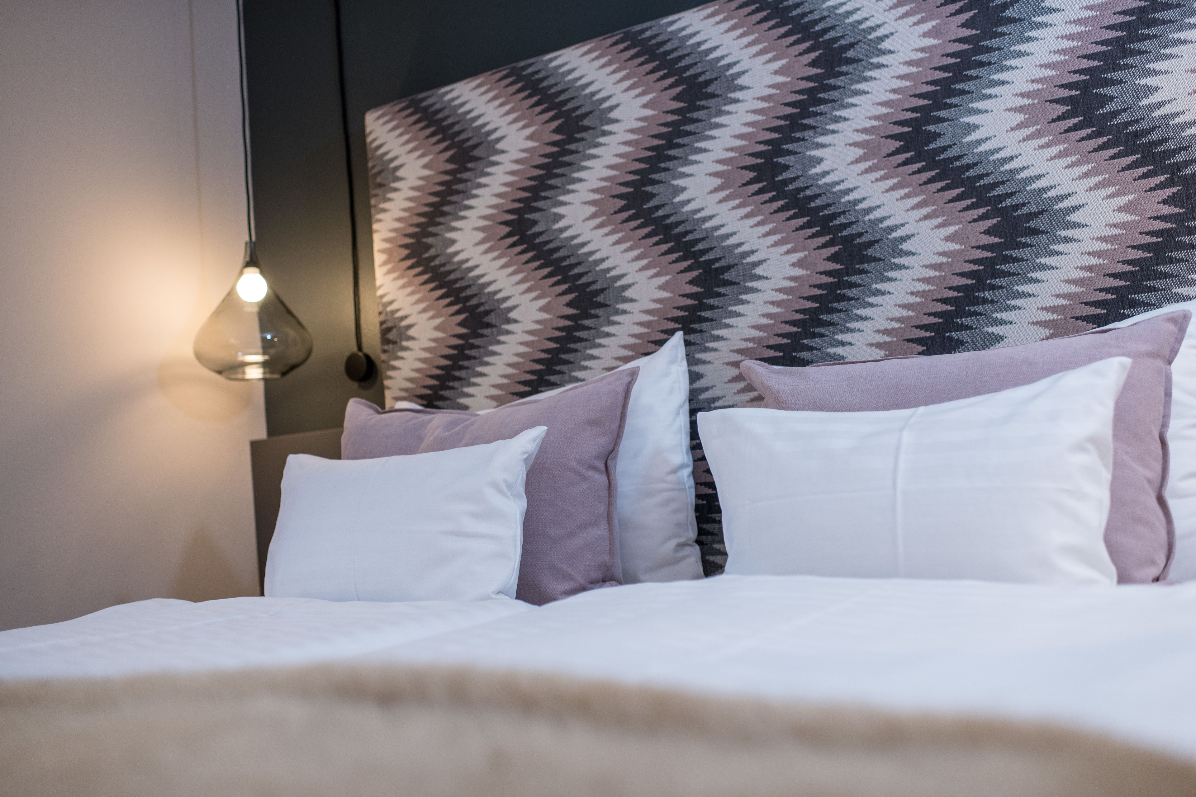 A bed with white and pink bedding, a patterned headboard, and a hanging light fixture.