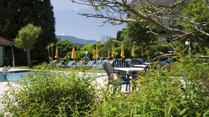 A poolside area with lounge chairs and umbrellas, surrounded by greenery and a view of mountains. - Kaiser Franz Josef (Millstatt)