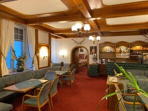 A cozy restaurant with wooden beams, green upholstered seating, and a bar area with shelves of bottles. - Kaiser Franz Josef (Millstatt)