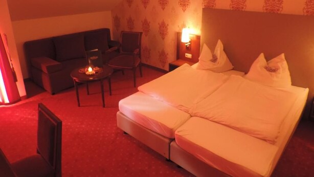 A hotel room with a bed, a sofa, a chair, a small table, and a nightstand with a lamp. - Kaiser Franz Josef (Millstatt)