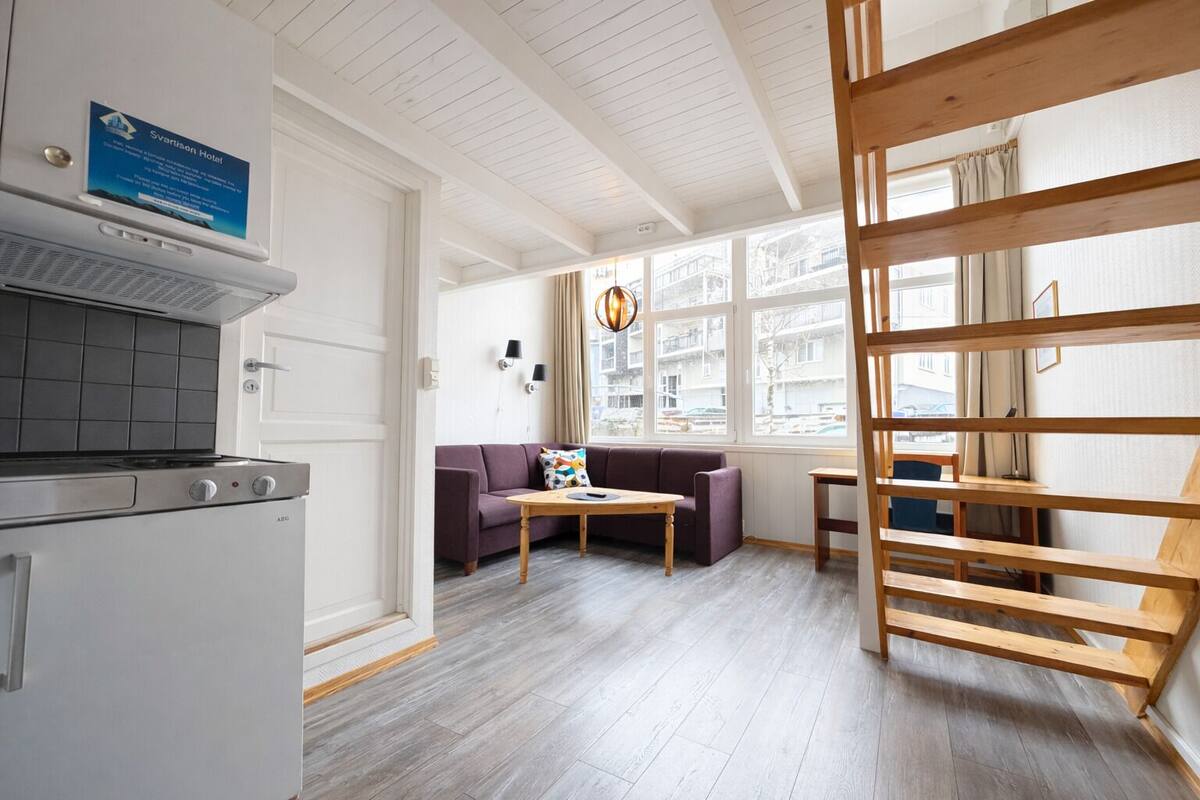 A modern kitchen with a built-in stove, a purple sofa, a wooden table, and a view of the city through large windows.
