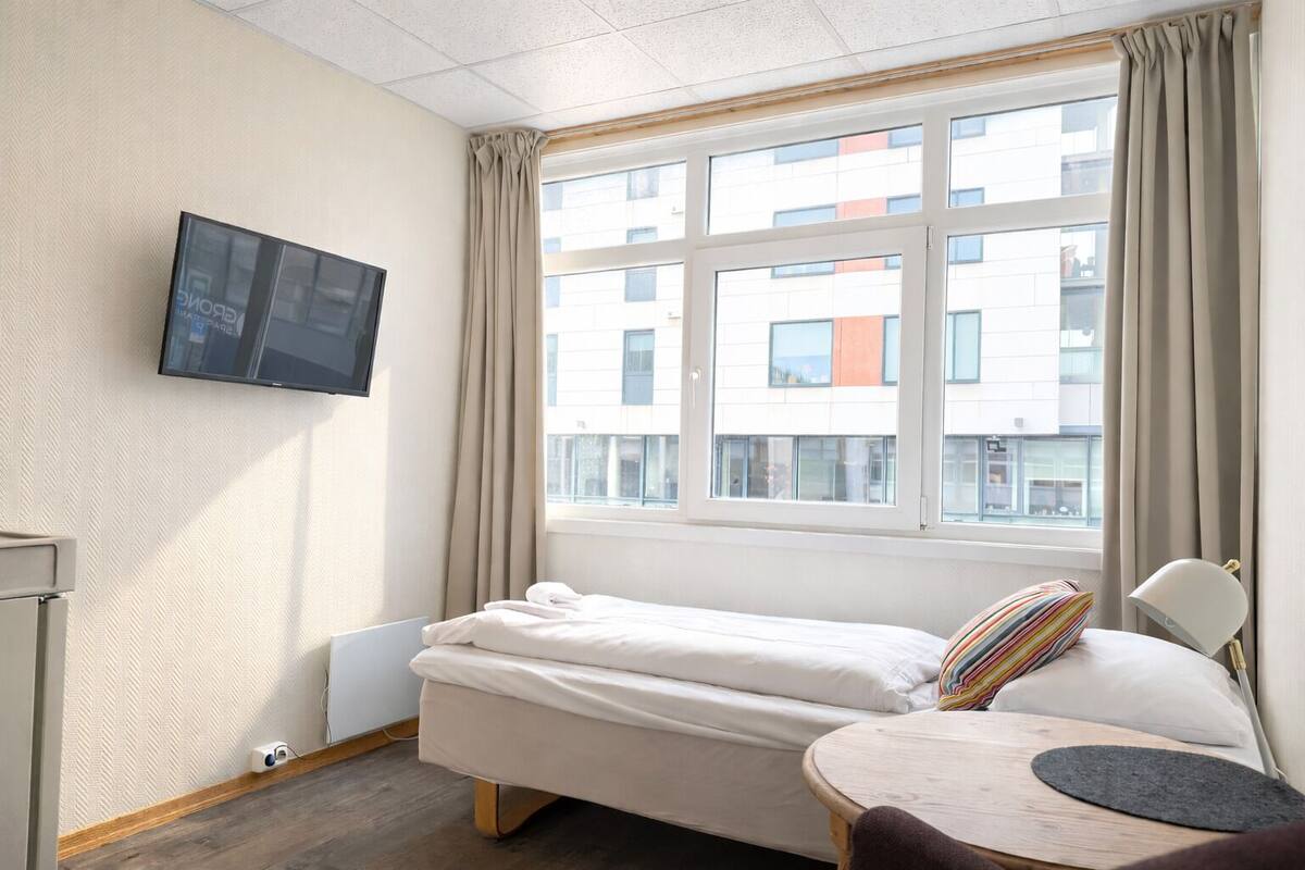 A compact hotel room with a bed, a small table, a chair, a TV mounted on the wall, and a window with curtains.