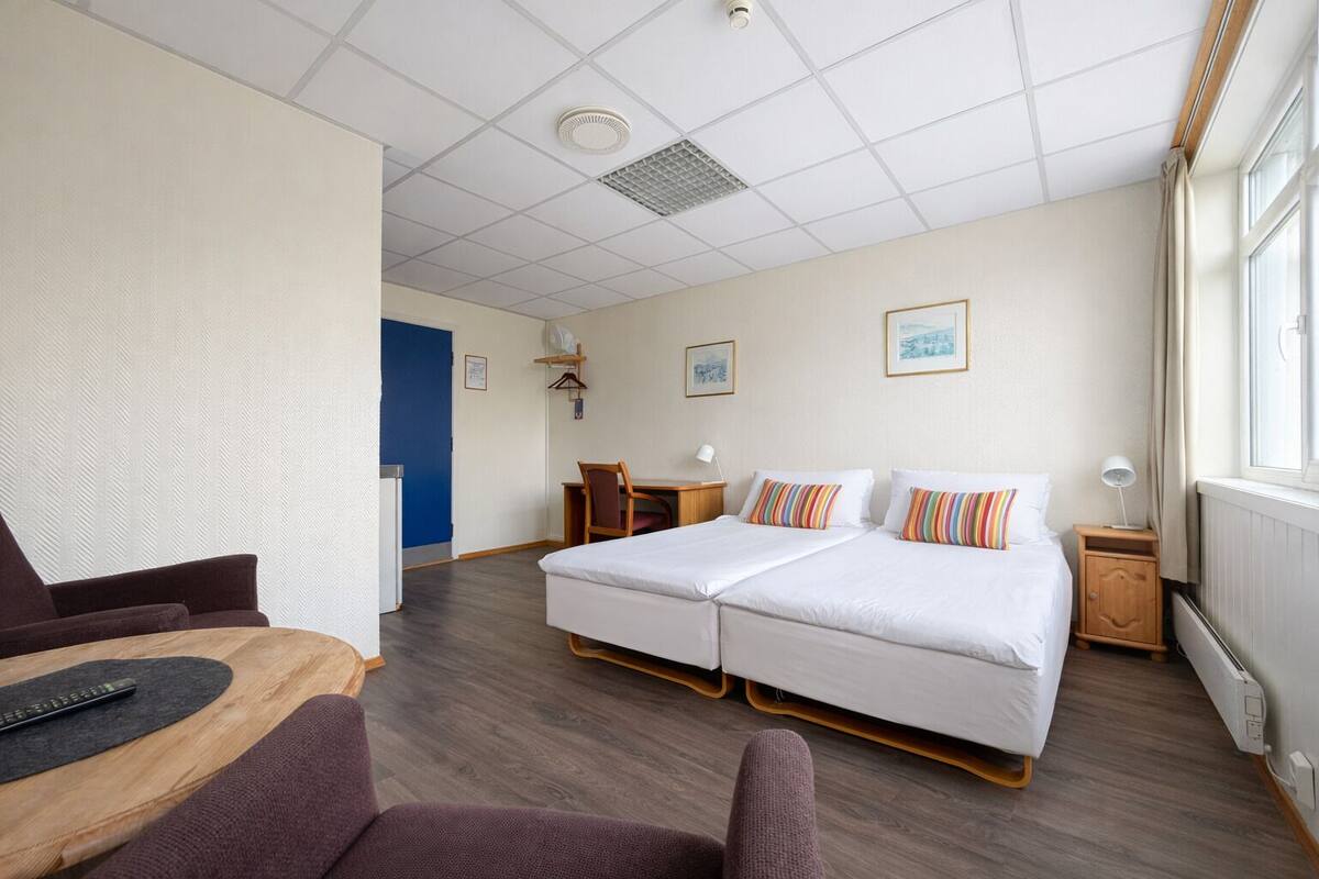 A hotel room with a double bed, a desk, a chair, a small table, a radiator, and a window.