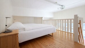 Ein modernes Schlafzimmer mit Holzboden, weißer Bettwäsche, einem Nachttisch mit Lampe und einem Balkon mit Geländer.