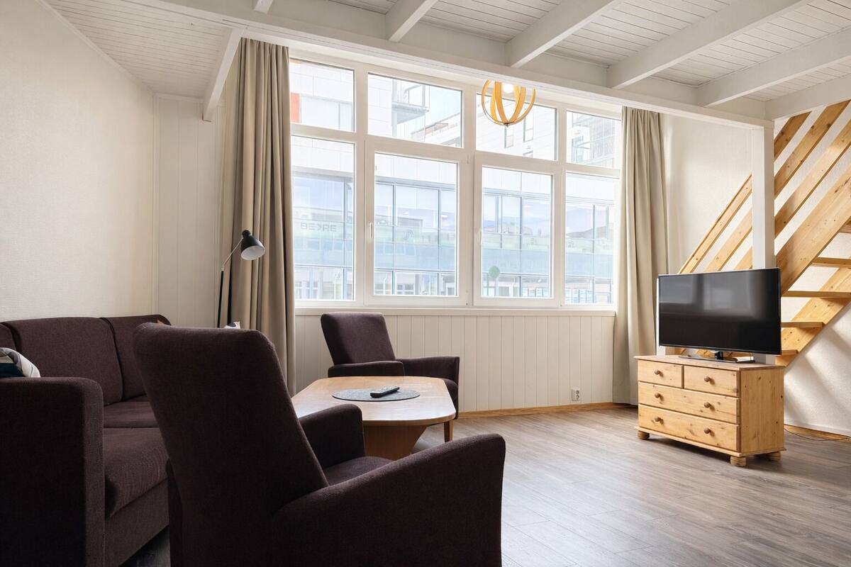 A modern living room with a sofa, chairs, a TV on a wooden cabinet, a table, and large windows with curtains.