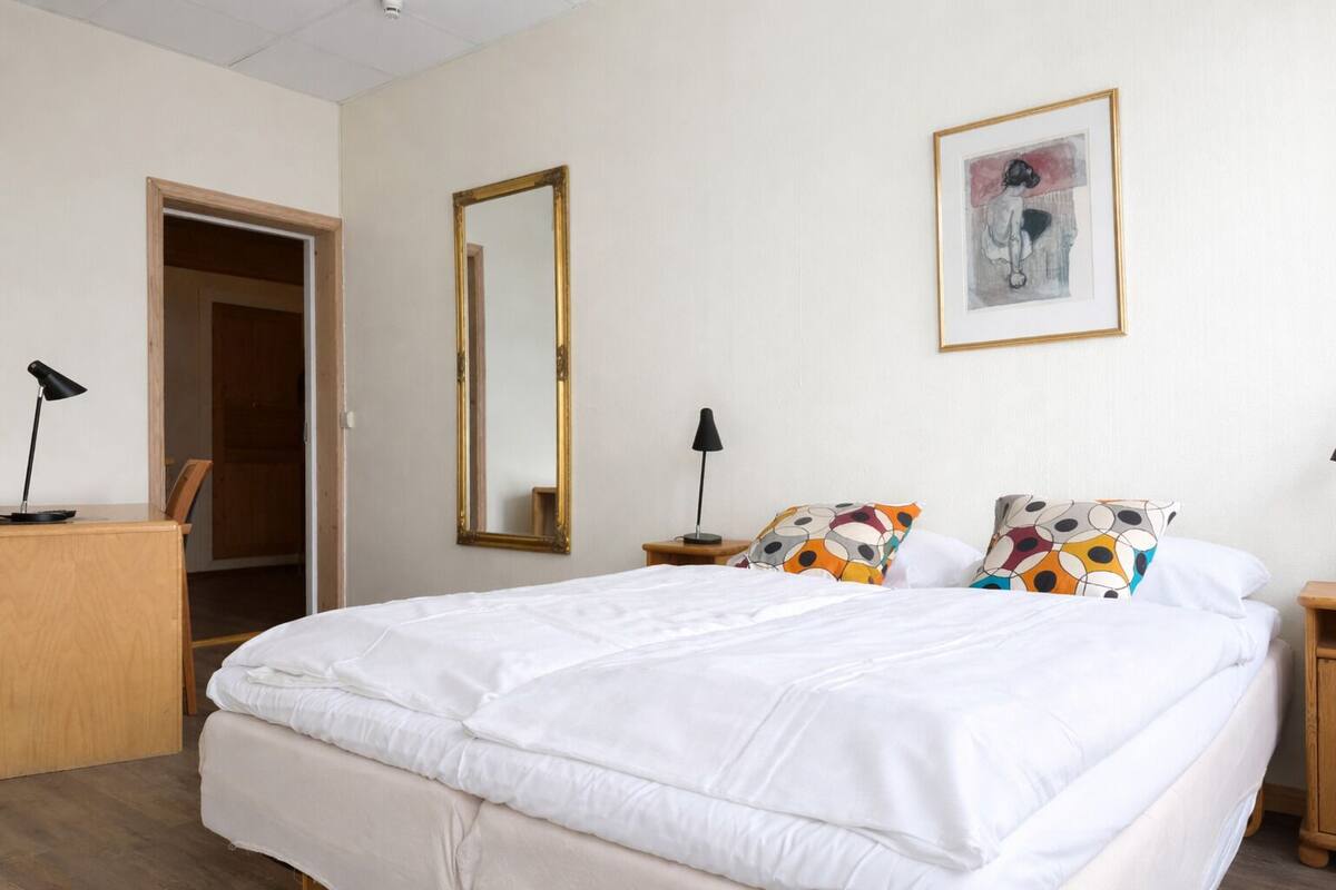 A bedroom with a large bed, a wooden desk, a mirror, a nightstand, and a framed picture on the wall.