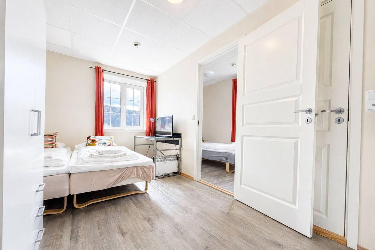 A hotel room with a bed, a desk with a computer, a window with curtains, and a door.
