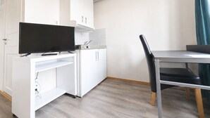 A compact kitchen with white cabinets, a black TV on a white stand, a small sink, and a dining table with a black chair.