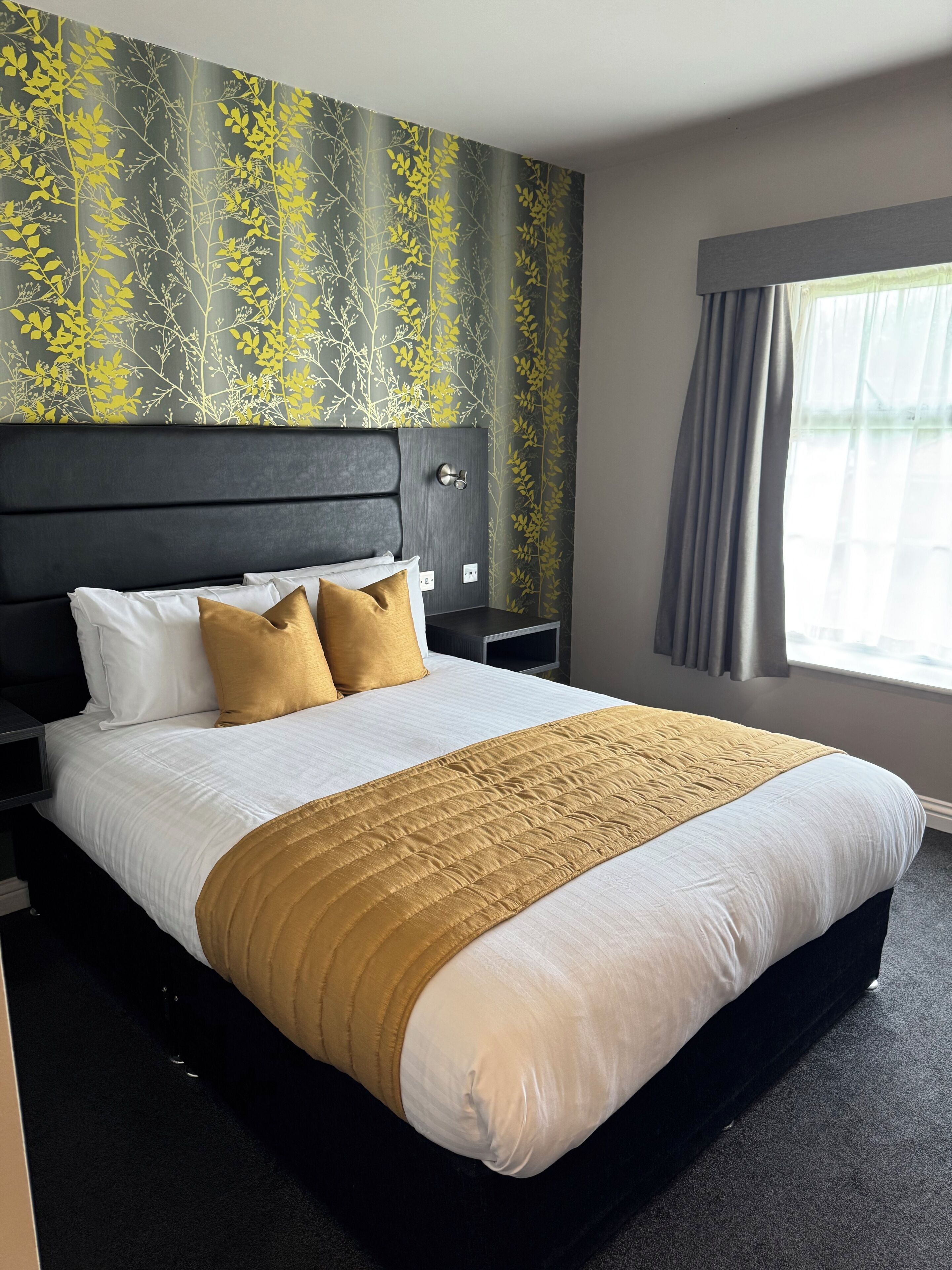 A hotel room with a large bed, a nightstand, a wall-mounted TV, and a window with curtains.