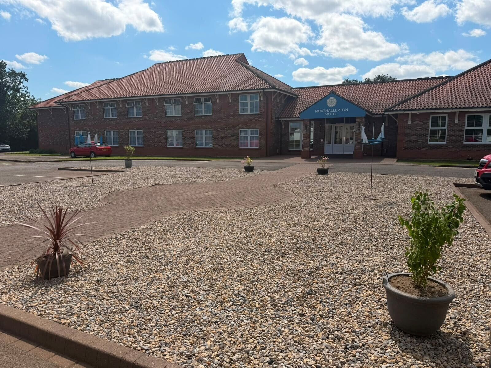 A brick building with a tiled roof, a sign reading 'Northallerton Motel,' and a gravel parking area with potted plants.