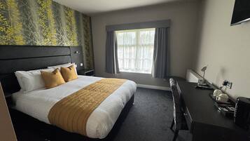 Ein Hotelzimmer mit Bett, Schreibtisch, Stuhl, einem Fenster mit Vorhängen und einem am Wand montierten Fernseher.