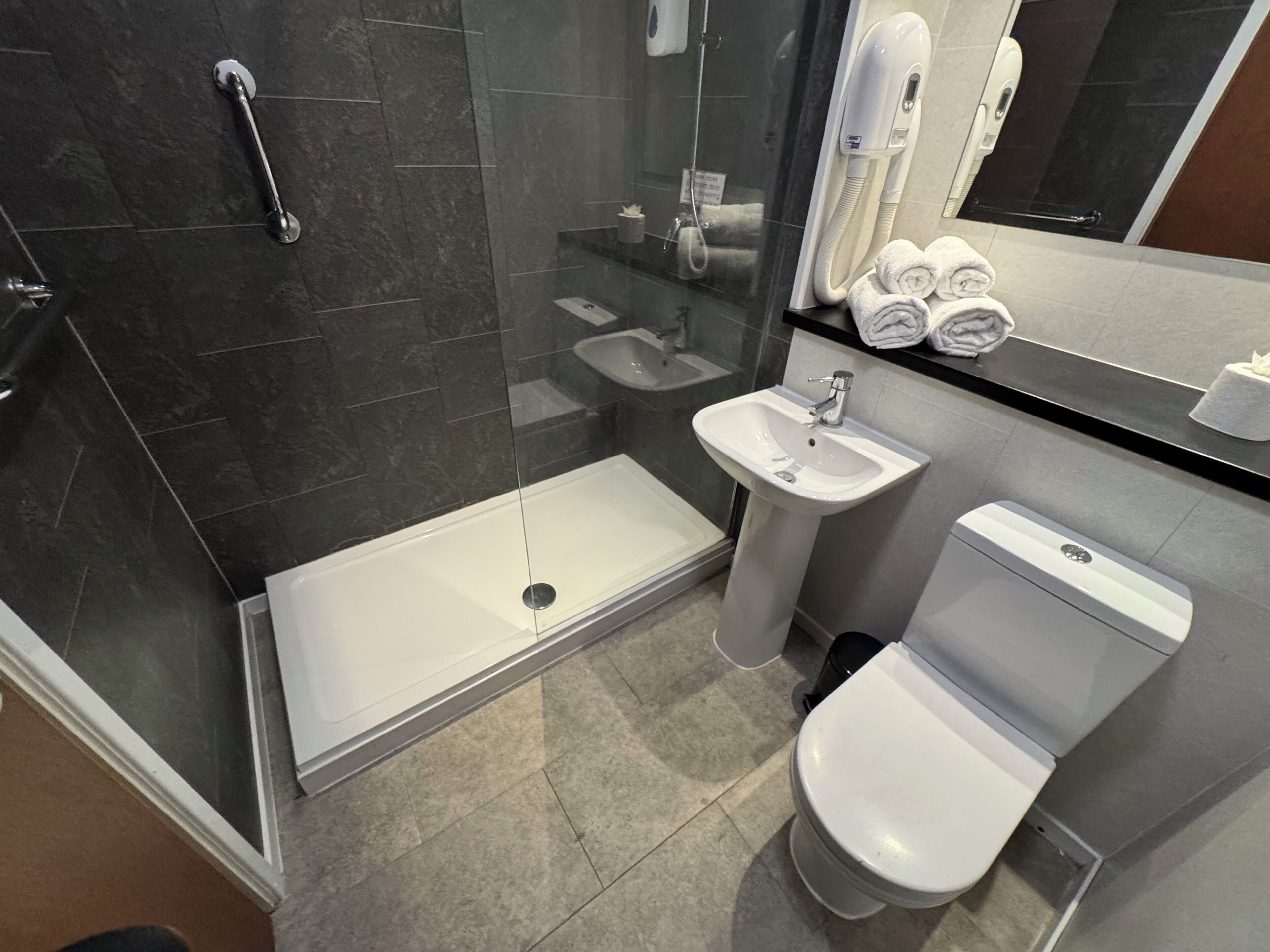 Un baño moderno con ducha envuelta en vidrio, lavamanos sobre pedestal, inodoro y espejo sobre el lavamanos.