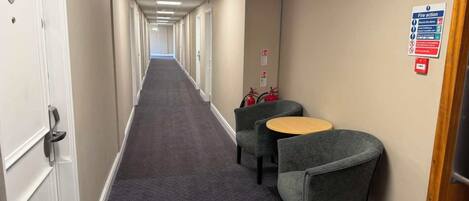 A hotel corridor with a carpeted floor, doors on the left, and a fire action sign on the right.