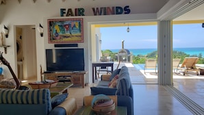 A living room with a TV, sofas, and a painting. It has a view of the ocean and a sign that reads "FAIR WINDS". - Villa Fair Winds (Jolly Harbour)