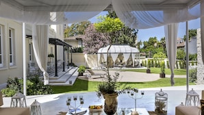 An outdoor patio with a table set for dining, surrounded by white drapes and a view of a garden and a gazebo. - Hotel Villa Fanny (Cagliari)