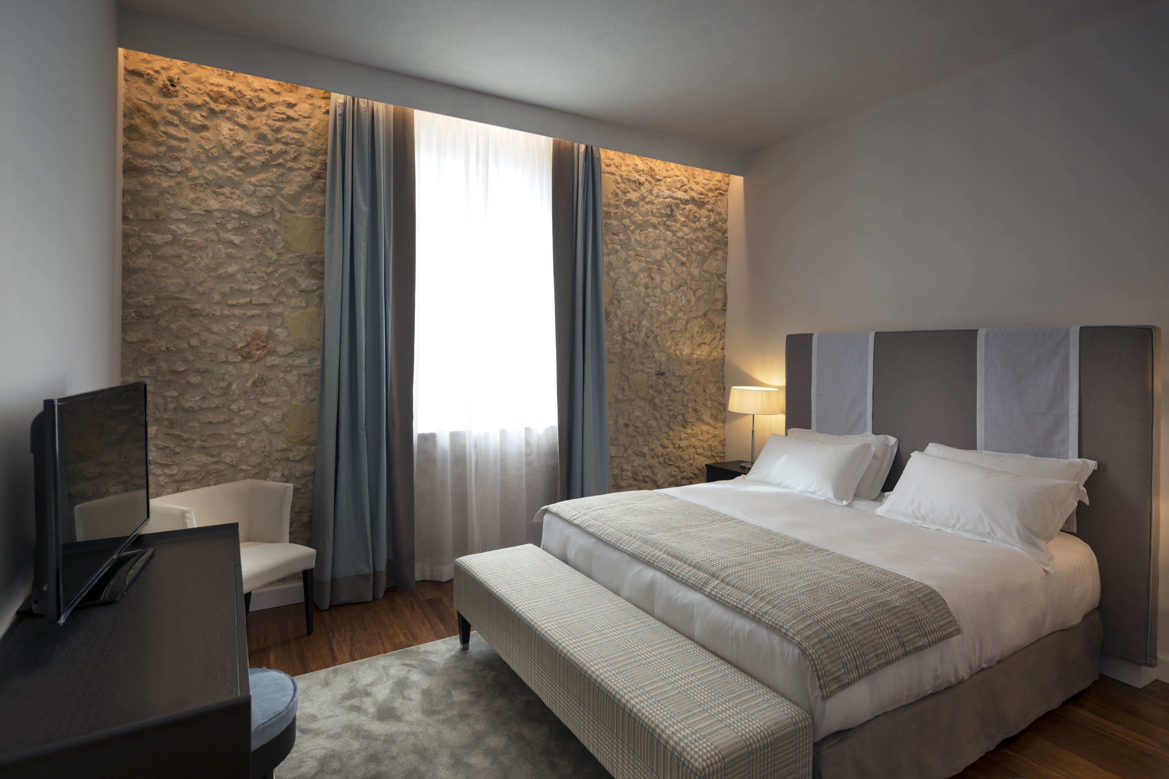 Habitación de hotel moderna con pared de acento de piedra, una cama grande con cabecera, una mesita de noche con lámpara, un televisor de pantalla plana, un banco y una ventana con cortinas.