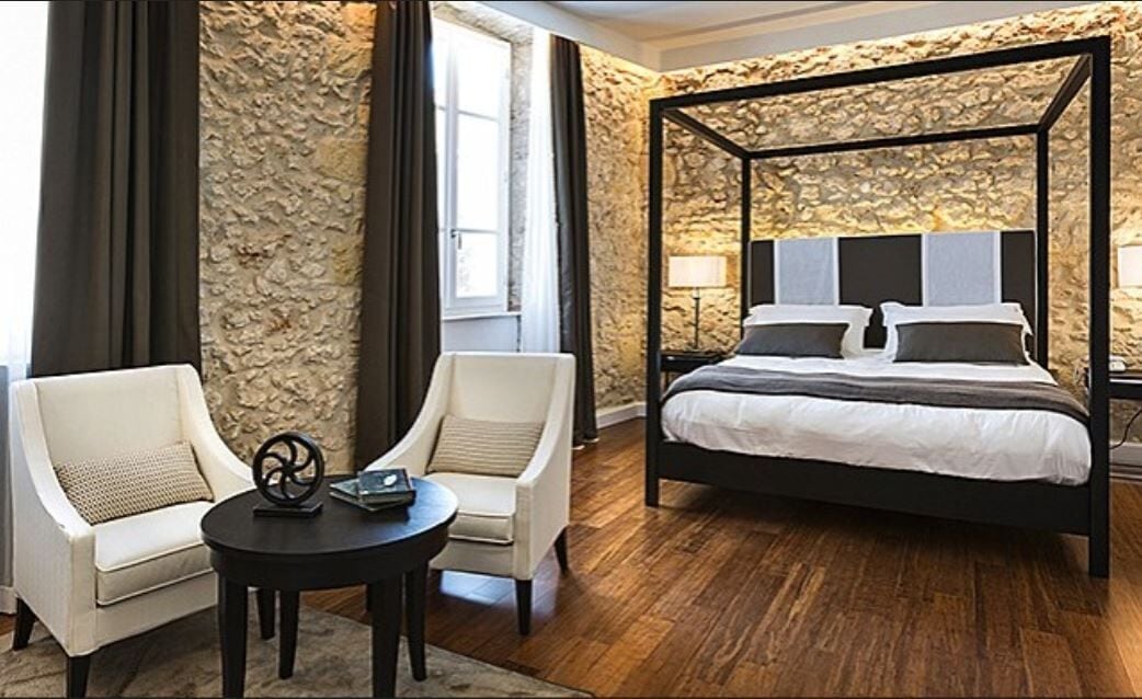 A bedroom with a four-poster bed, two armchairs, a round table, and a stone wall.