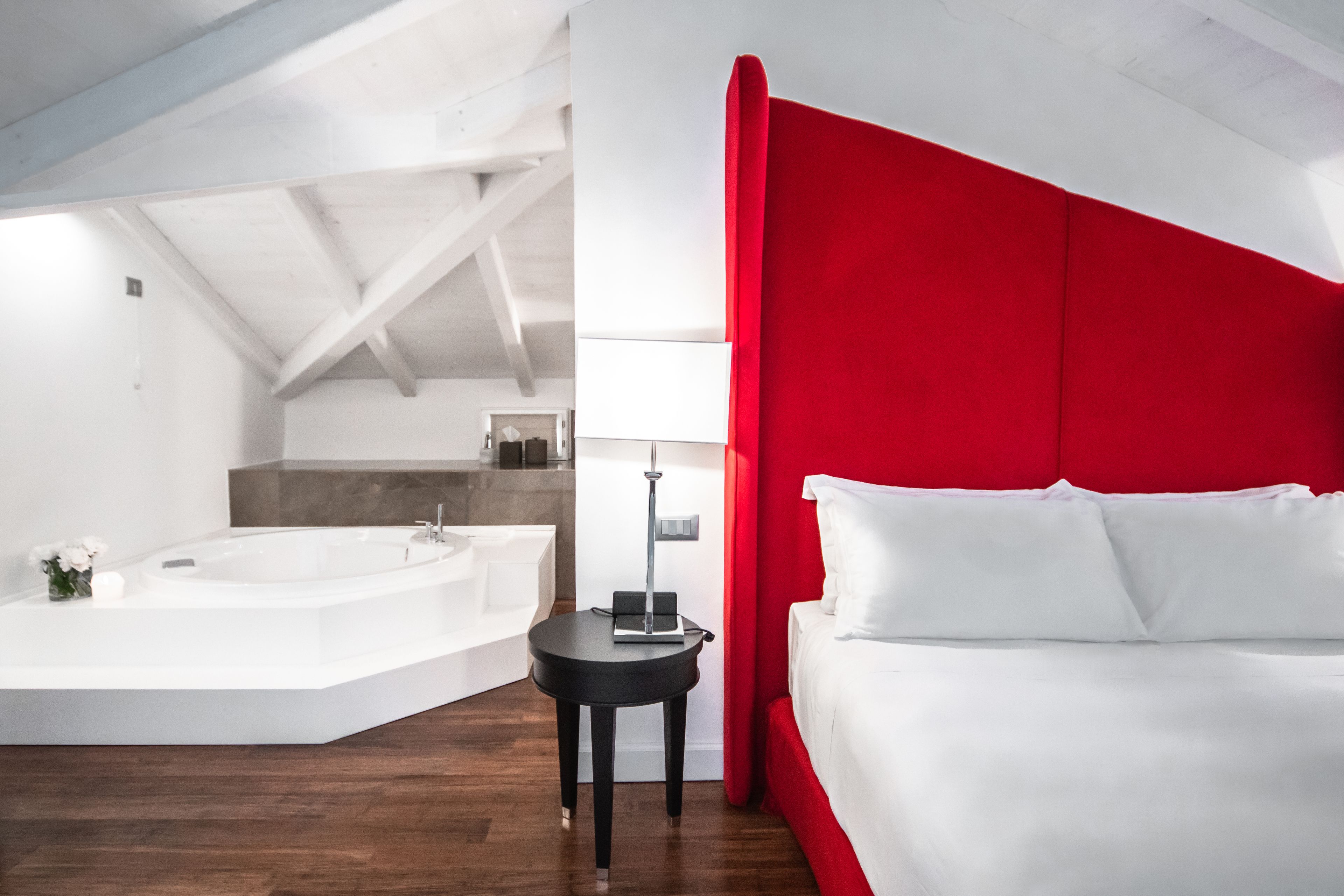 Ein modernes Hotelzimmer mit einem großen Bett, einer roten Akzentwand, einem separaten Badezimmer mit einer Badewanne und einem kleinen Tisch mit einer Lampe.