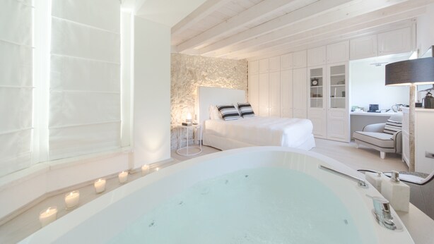 A modern bedroom with a large bathtub, a bed with white bedding, and a stone accent wall. - Hotel Villa Fanny (Cagliari)