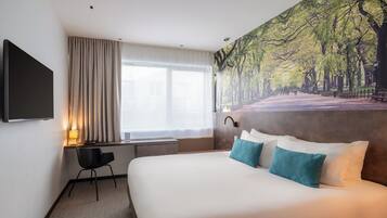 Habitación de hotel con una cama grande, un escritorio con silla, un televisor montado en la pared y un ventanal con cortinas.