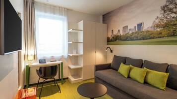 Une chambre d’hôtel moderne équipée d’un bureau, d’une chaise, d’un canapé et ornée d’un tableau représentant un paysage urbain.