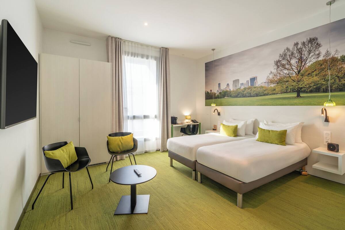 A hotel room with two beds, a desk, a chair, a remote control, and a painting of a cityscape.