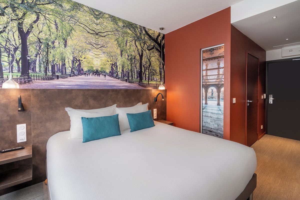 A hotel room with a large bed, a mural of a park, a door, and a small table with a lamp.