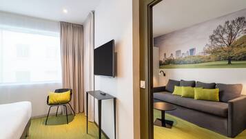 Ein Hotelzimmer mit einem Bett, einem Sessel mit gelbem Kissen, einem kleinen Tisch, einer grauen Couch mit grünen Kissen, einem an der Wand montierten Fernseher und einem Gemälde, das einen Park mit Bäumen und eine Stadtansicht zeigt.