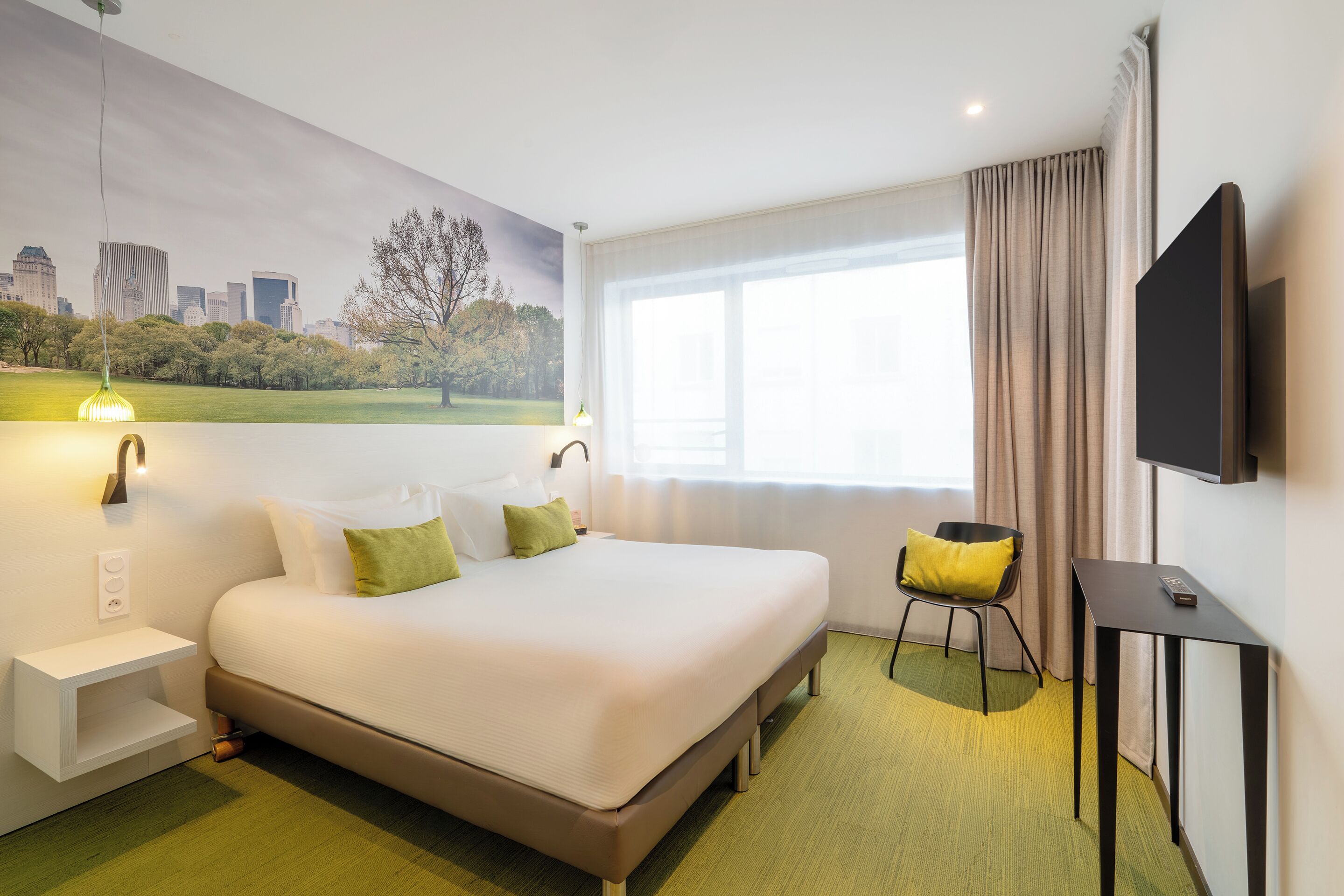 Una habitación de hotel con cama, una silla, televisor, ventana con cortinas, lámpara de pared y un cuadro de un paisaje urbano.