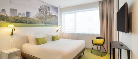 A hotel room with a bed, a chair, a TV, a window with curtains, a wall-mounted light, and a painting of a cityscape.