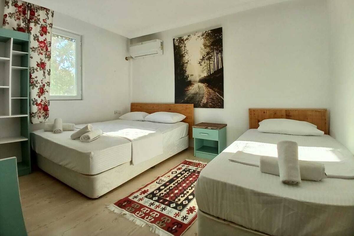 A room with two single beds, a small table, a shelf, a window with floral curtains, and a poster on the wall.