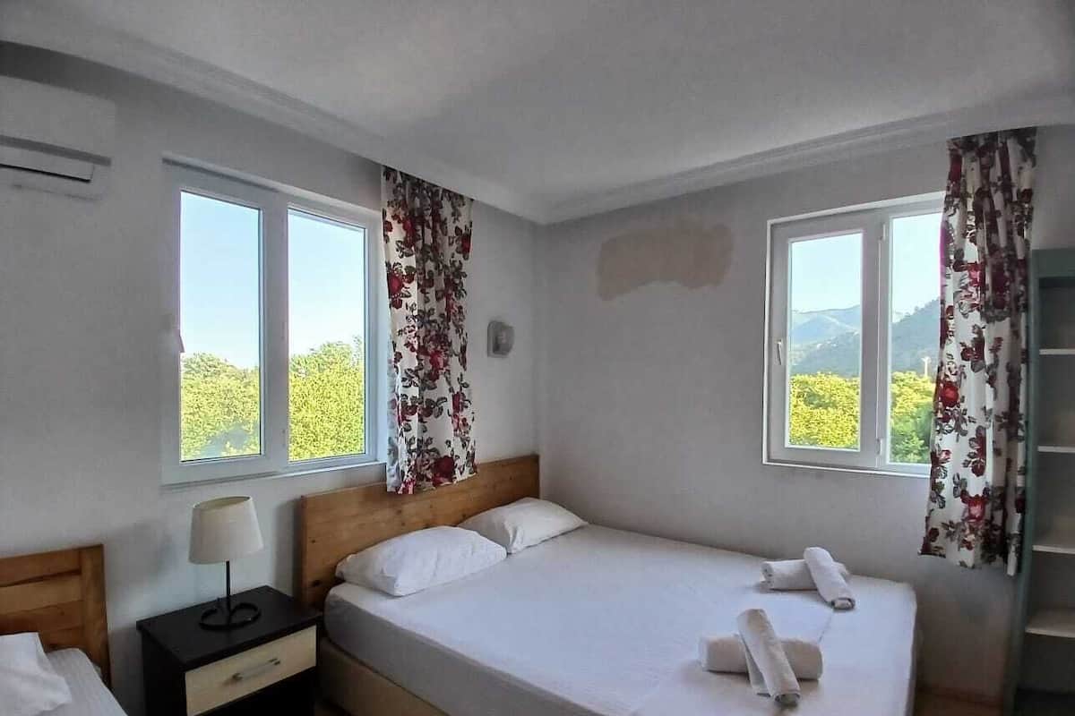 A single-bed room with a wooden headboard, a bedside table with a lamp, and two windows with floral curtains.