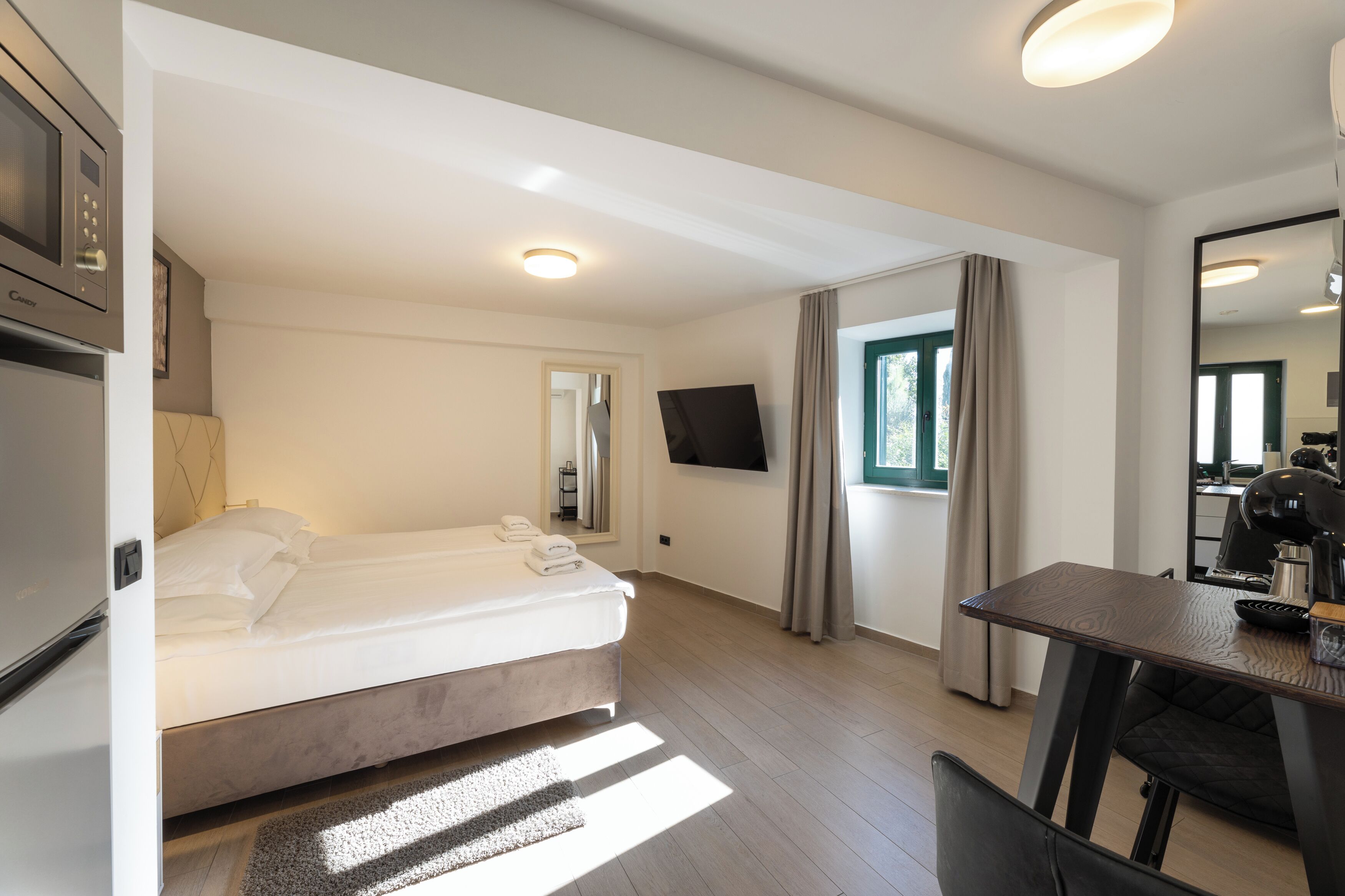 A modern hotel room with a bed, a dining table, a TV, and a kitchenette.
