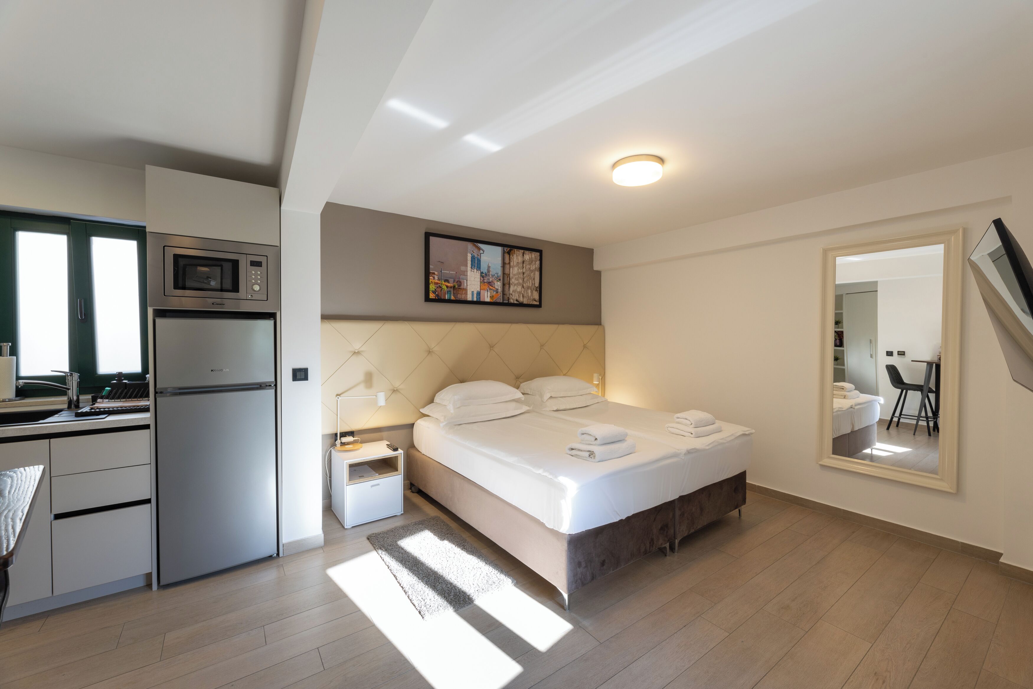 Una habitación de hotel moderna con cama, microondas integrado, una pequeña kitchenette y un espejo grande.