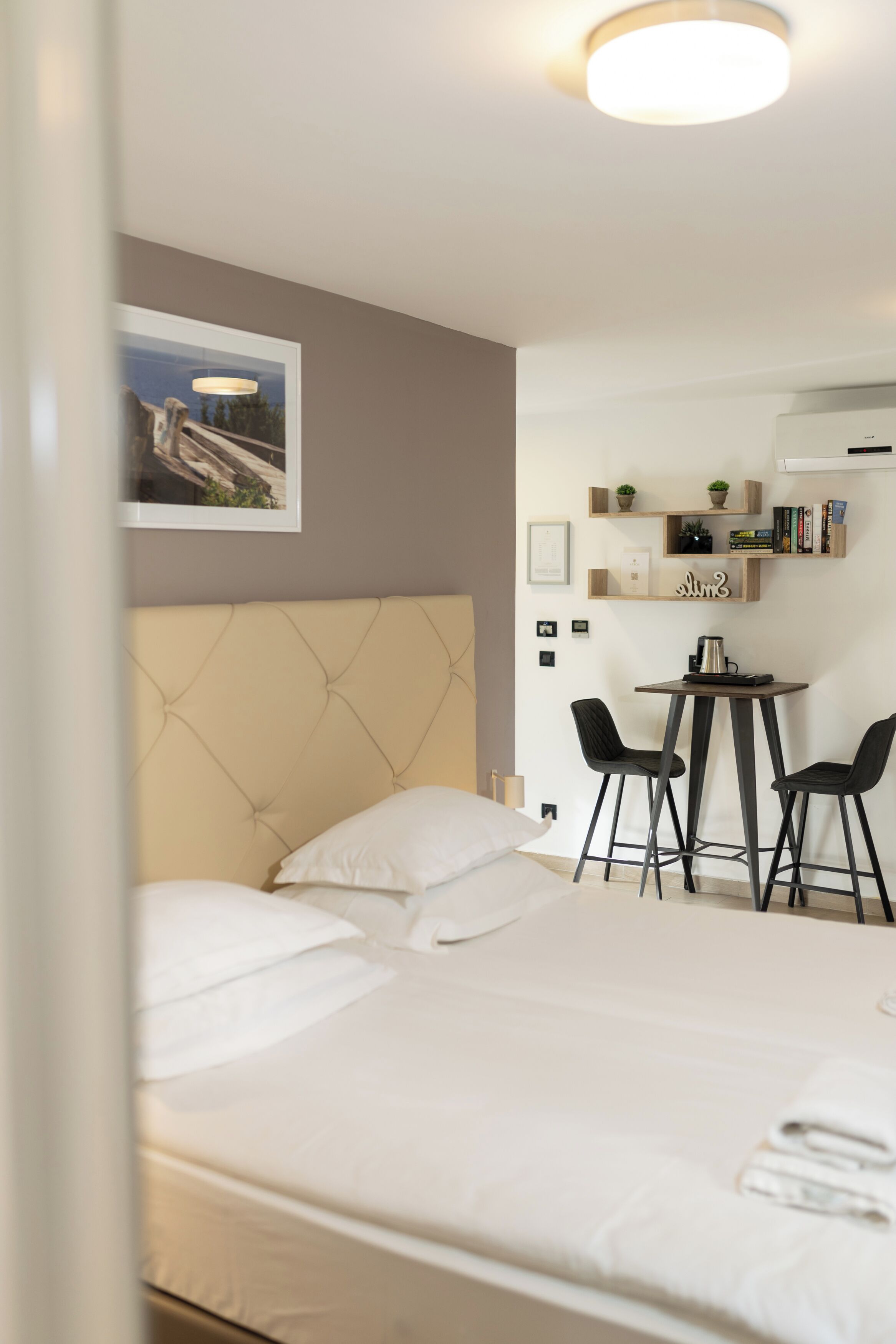 A hotel room with a bed, a small table with two chairs, a ceiling light, and a wall-mounted shelf with books and a plant.